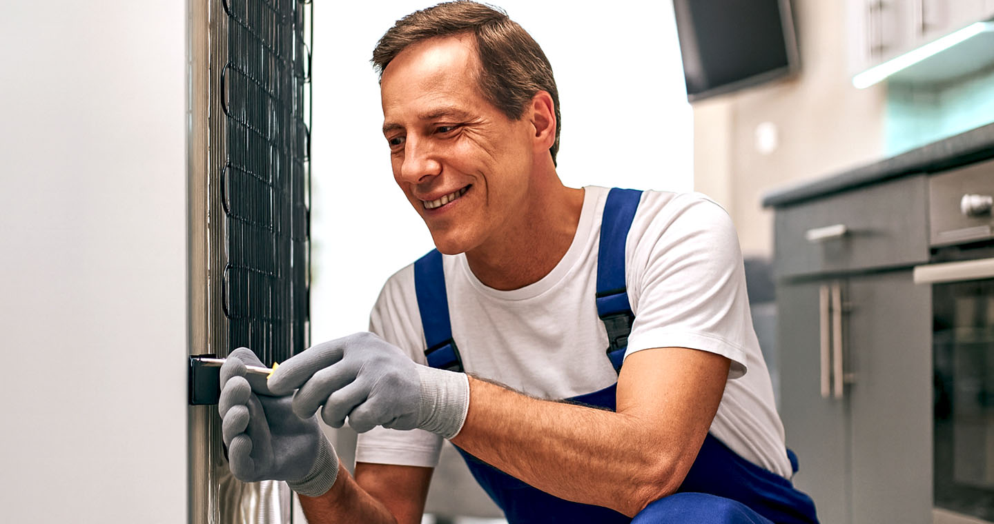 Smiling technician in blue overalls repairs the back of a refrigerator in a modern kitchen